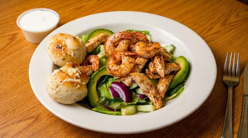 A vibrant salad with grilled shrimp, chicken, avocado, red onion, and garlic knots on a white plate.