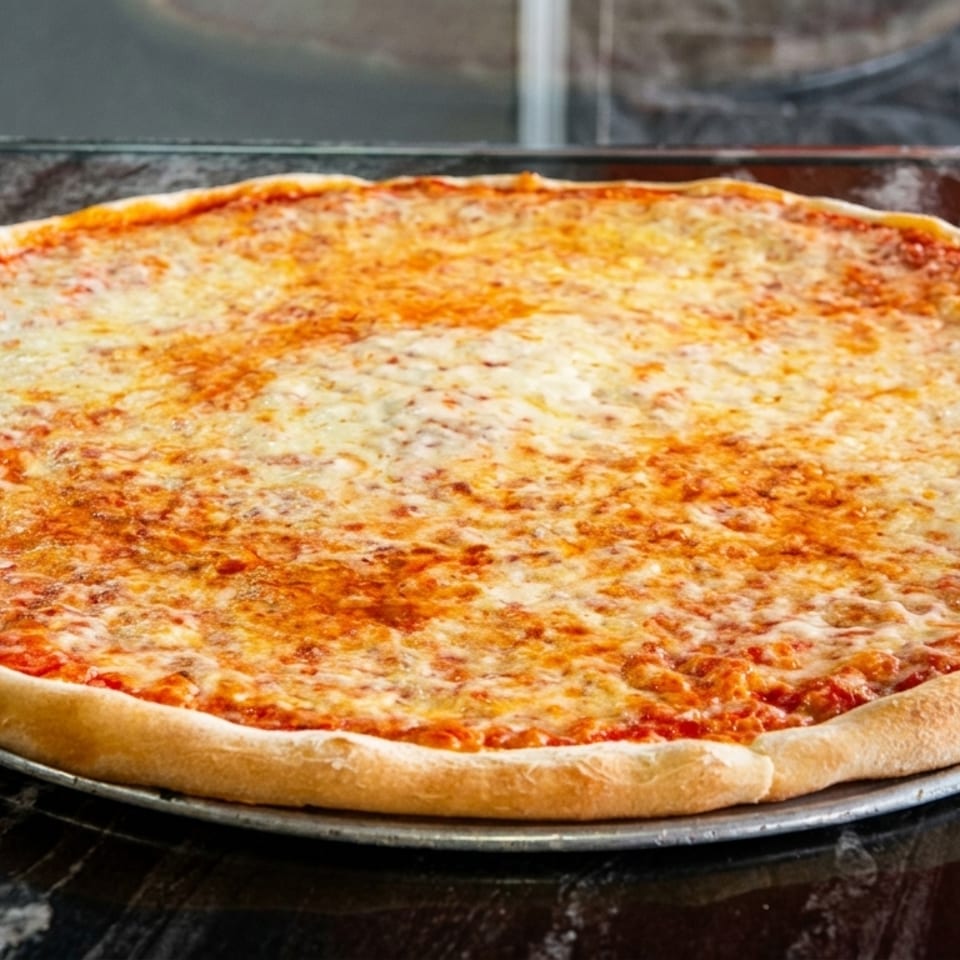 A large, round cheese pizza with a golden crust sits on a dark, reflective counter.