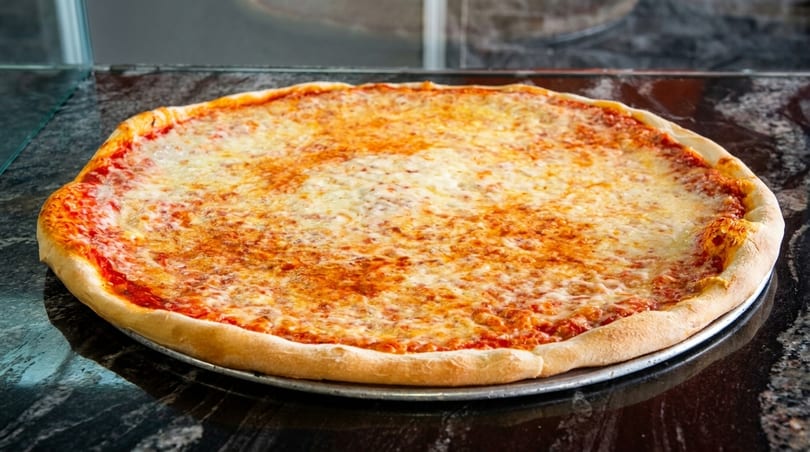 A large, round cheese pizza with a golden crust sits on a dark, reflective counter.